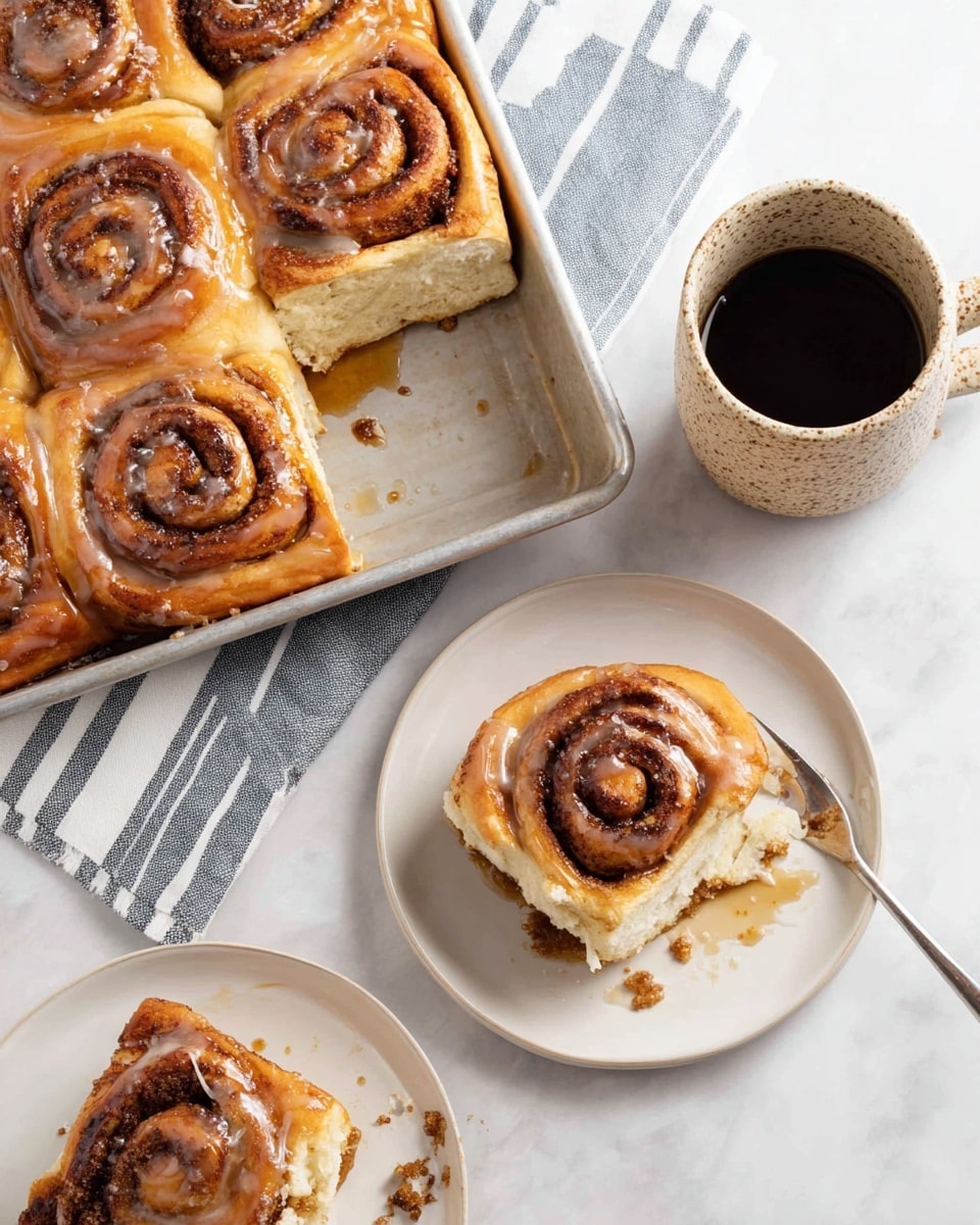 The image shows a tray of cinnamon rolls with a golden brown, glossy top layer glazed with a light syrup. Each roll has visible dark brown cinnamon swirls tightly spiraled in soft, fluffy dough with a creamy light beige center. Two rolls are served on separate white plates; one is cut showing the soft inner layers, and both have syrup drizzled on them with some cinnamony crumbs around. A speckled beige ceramic mug filled with black coffee is placed near the plates, all set on a white marbled textured surface with a folded striped cloth under the baking tray. photo taken with an iphone --ar 4:5 --v 7
