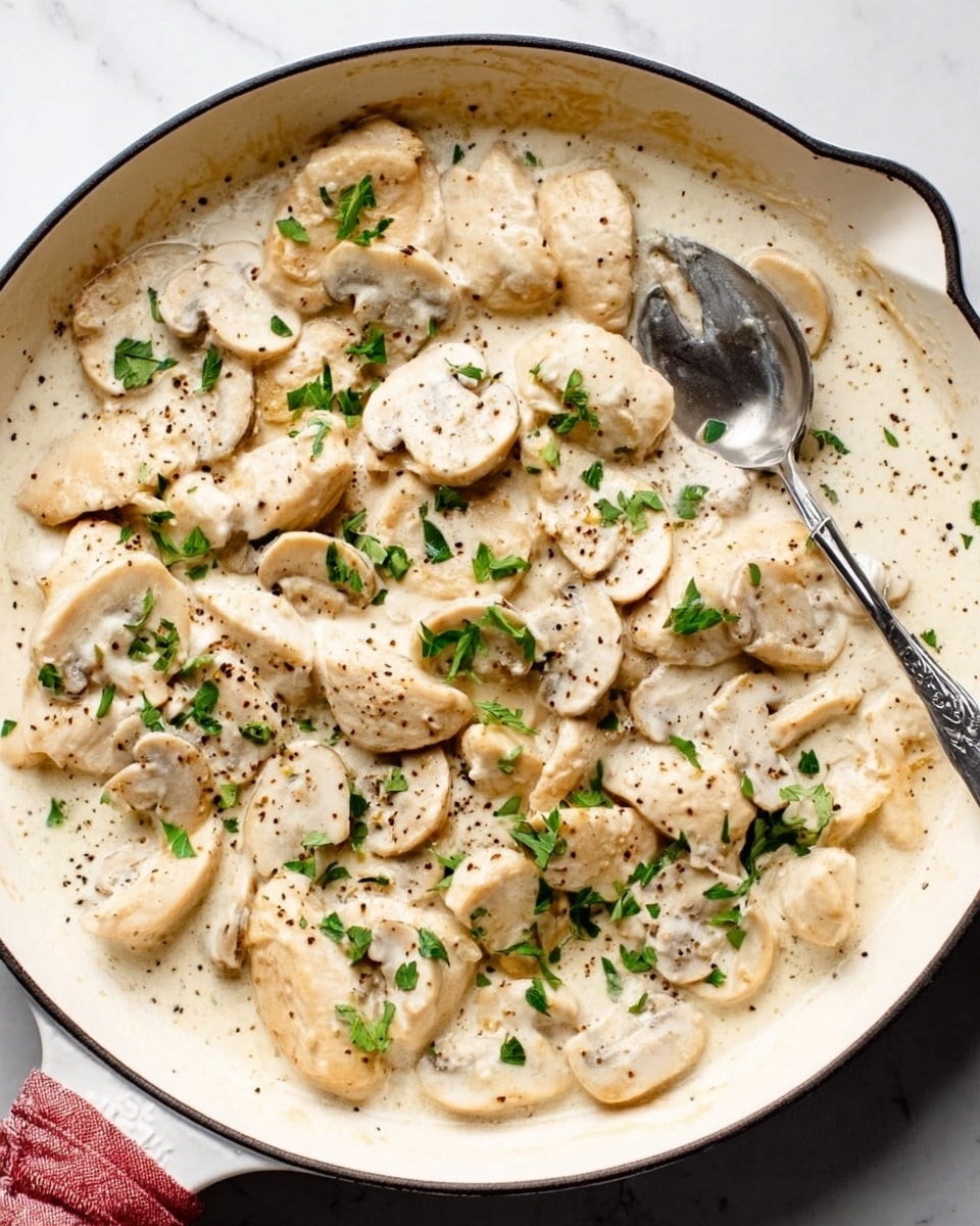 A white skillet with a creamy white sauce filled with many slices of light beige chicken and white mushrooms, sprinkled with small pieces of green parsley and black pepper on top, a silver spoon rests inside the skillet. The skillet is placed on a white marbled surface. photo taken with an iphone --ar 4:5 --v 7