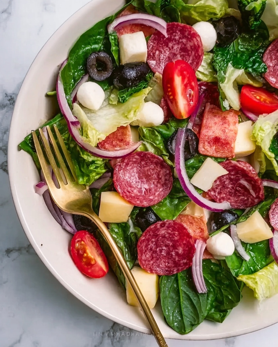 A large white plate holds a fresh salad with multiple layers: the base is dark green spinach and lighter green lettuce leaves, layered with bright red cherry tomato halves and thin purple onion slices. Scattered among the greens are black olive slices, cubes of pale yellow cheese, and small white mozzarella balls. Thin slices of red and pink salami are laid on top, adding texture and color contrast. A gold fork rests on the plate's edge, positioned over the salad. The background is a white marbled surface. Photo taken with an iphone --ar 4:5 --v 7