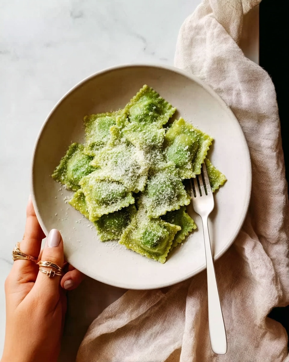 A white plate sits on a white marbled surface with a soft beige cloth nearby. On the plate is a single layer of green ravioli pasta, arranged loosely in the center. The ravioli have a slightly rough texture and are topped with a light dusting of grated white cheese. A woman's hand with rings on her fingers holds a white fork gently touching the edge of the plate. The overall look is simple and fresh, with a natural light highlighting the colors and textures. photo taken with an iphone --ar 4:5 --v 7