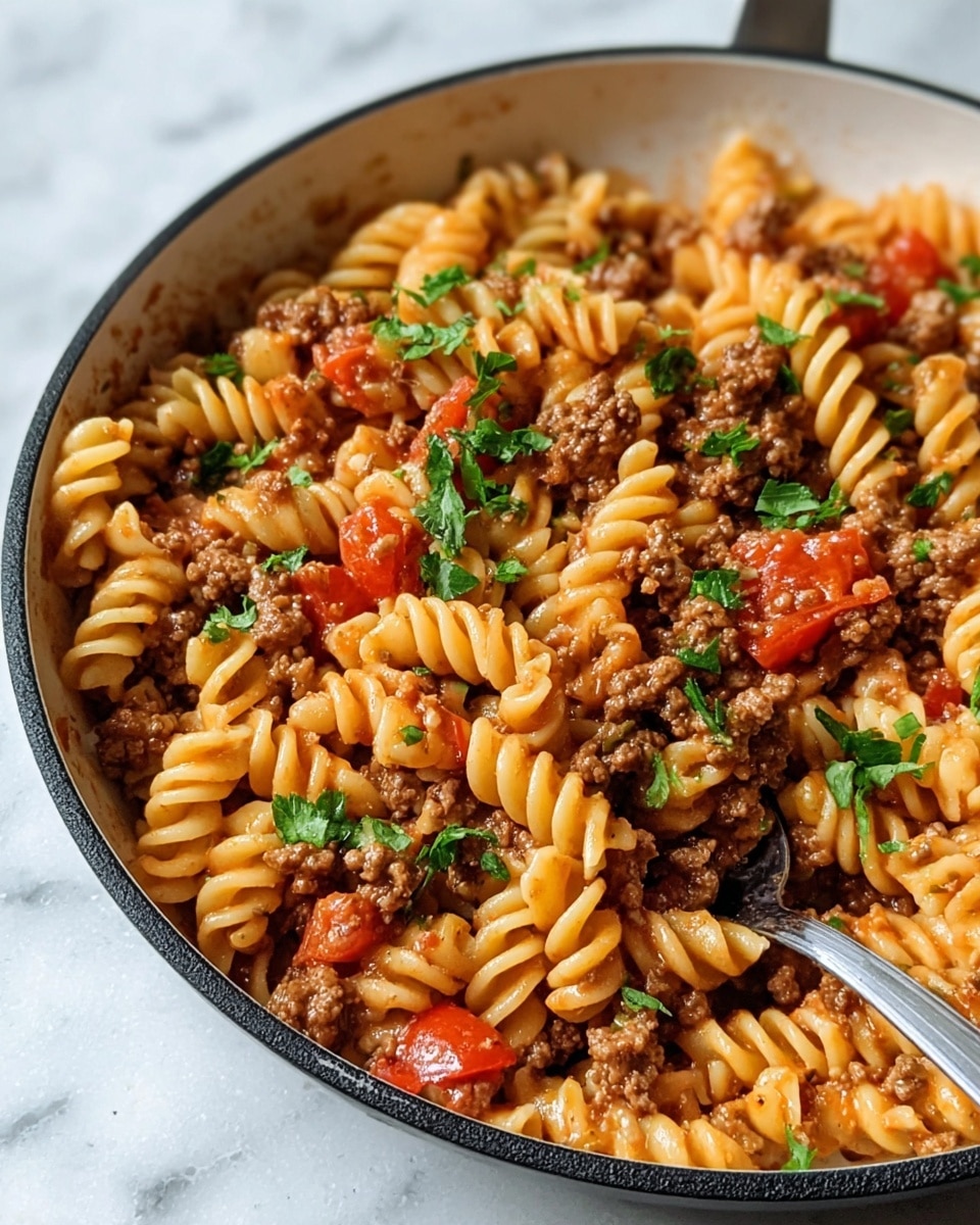The image shows a close-up of a skillet filled with spiral pasta mixed with cooked ground meat and tomato chunks. The pasta is light orange with a smooth texture, twisted in spirals. The ground meat is brown and crumbly, evenly spread among the pasta. Bright red tomato pieces add color and moistness. Fresh chopped green herbs are sprinkled on top, adding a touch of freshness. The skillet is white with a black rim, sitting on a white marbled background. A spoon is partially visible on the right side inside the skillet. Photo taken with an iphone --ar 4:5 --v 7