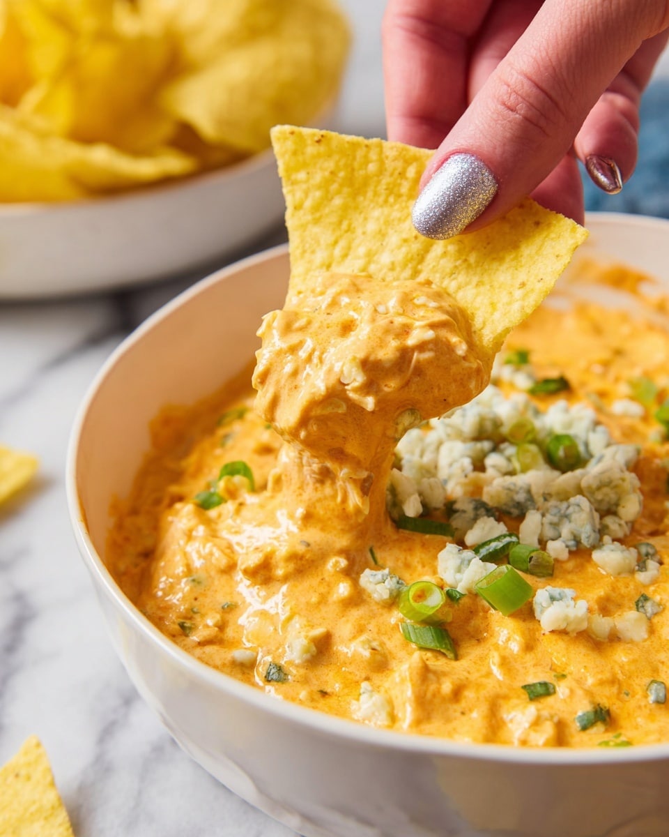 A close-up of a yellow corn chip being dipped into a creamy orange cheese dip with a thick texture, sprinkled with small green onion pieces and chunks of blue cheese on top; the dip is served in a white bowl, held by a woman's hand with silver-painted nails, all set on a white marbled surface with another white bowl of yellow chips blurred in the background. Photo taken with an iphone --ar 4:5 --v 7