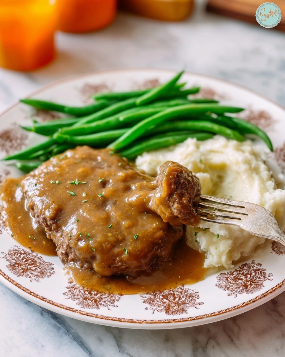 A white plate with a brown floral pattern holds a meal of three layers: on the left, bright green cooked green beans with smooth texture are stacked close together; on the right, a pile of creamy white mashed potatoes forms a soft base; covering the potatoes and part of the plate are two round beef patties in the middle, smothered in thick, rich brown gravy with visible herbs sprinkled on top. A fork in the front right pierces a piece of the beef patty covered with gravy, lifting it slightly above the plate. The plate sits on a white marbled surface with some orange blurred objects in the background. Photo taken with an iphone --ar 4:5 --v 7
