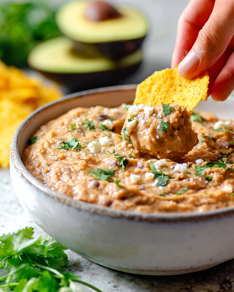 A close-up view of a white bowl filled with creamy light brown refried beans mixed with small specks of white cheese and sprinkled green cilantro leaves, showing a thick texture; in the foreground, a yellow corn chip held by a woman's hand is dipped into the beans, capturing the creamy mixture clinging to the chip; the background features out-of-focus halves of green avocados and blurred cilantro on a white marbled surface. photo taken with an iphone --ar 4:5 --v 7