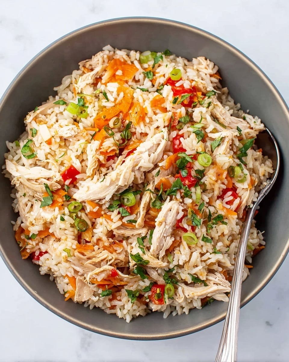 A grey bowl filled with a mix of cooked white rice, shredded light brown chicken pieces, and sliced bright orange carrots. Scattered green herbs and sliced green onions are sprinkled on top, adding fresh green color. There are pieces of red peppers mixed throughout, providing a pop of red color. A silver spoon rests inside the bowl on the right side. The bowl sits on a white marbled surface. photo taken with an iphone --ar 4:5 --v 7