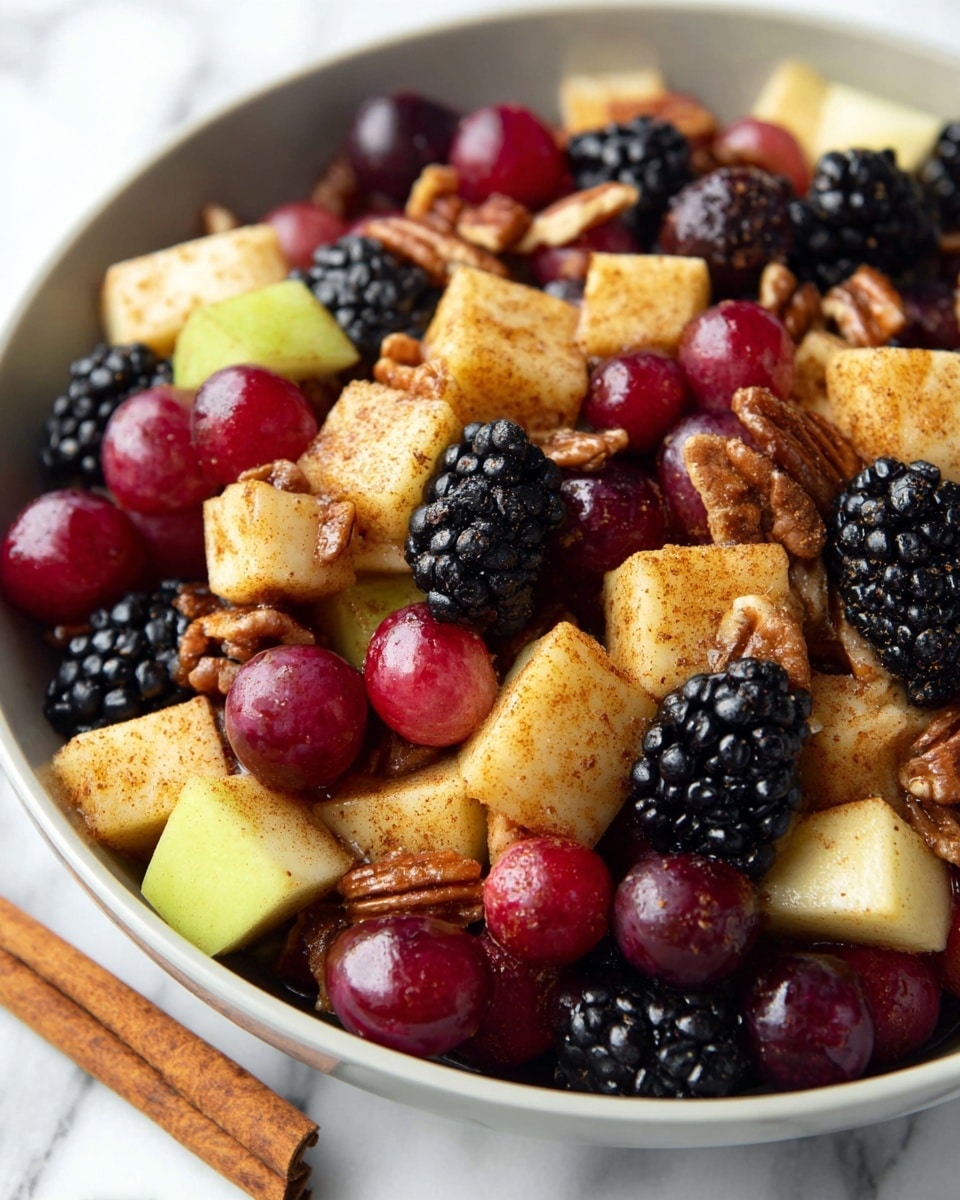 The image shows a close-up of a white bowl filled with a colorful fruit salad. The salad has three main layers: large apple cubes with a light brown cinnamon coating and a slightly rough texture, round shiny red grapes, and clusters of black, shiny blackberries. Mixed between these fruits are crunchy pecan halves with a textured brown surface. The bowl sits on a white marbled surface with two cinnamon sticks placed near it. The colors range from pale yellow of the apples, deep red of the grapes, dark black of the blackberries, and warm brown of the pecans, making the salad visually rich and appealing. Photo taken with an iphone --ar 4:5 --v 7