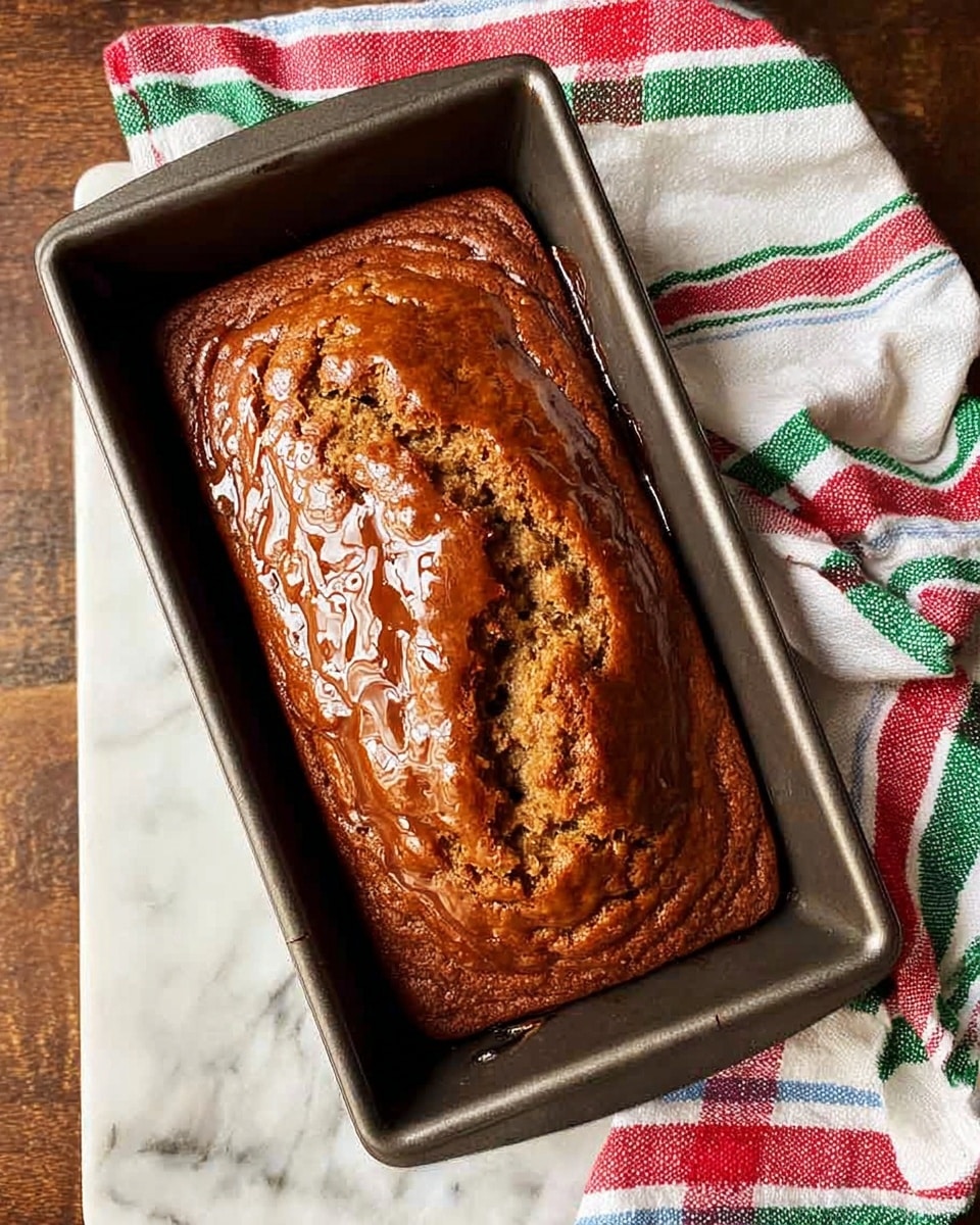 A rectangular loaf of banana bread sits inside a dark metallic baking pan placed on a white marbled surface. The bread has a rich golden-brown top with visible cracks and a shiny, sticky glaze covering the surface, pooling slightly at the edges. Next to the pan, a colorful white cloth with red, green, and blue stripes is casually draped, adding a cozy feel to the scene. photo taken with an iphone --ar 4:5 --v 7