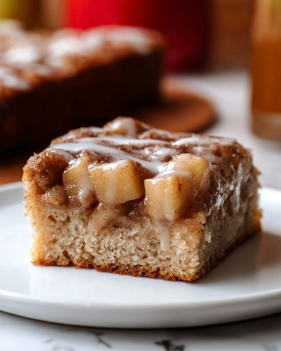 A close-up of a single square piece of cinnamon apple cake resting on a white plate over a white marbled surface; the cake has three visible layers—the bottom layer is a moist, light brown sponge, the middle layer is filled with soft, diced apple pieces coated in cinnamon sugar, and the top layer features a glistening, slightly translucent white glaze drizzled unevenly over the cinnamon-apple topping. In the background, blurred objects and a red surface add depth to the scene. photo taken with an iphone --ar 4:5 --v 7