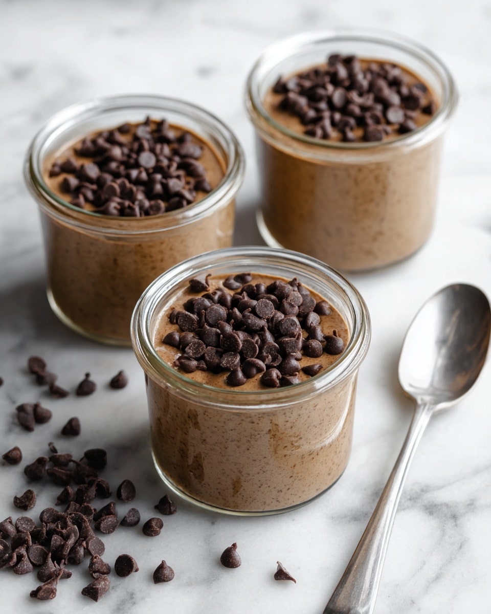 The image shows three glass jars filled with a creamy chocolate chia pudding, with visible small black chia seeds evenly spread throughout the light brown chocolate mixture. Each jar is topped with a layer of shiny dark chocolate chips, slightly piled in the center. The jars are placed on a white marbled surface with scattered chocolate chips around them, and soft natural light coming from the side highlights the texture of the pudding and the glossiness of the chocolate chips. photo taken with an iphone --ar 4:5 --v 7