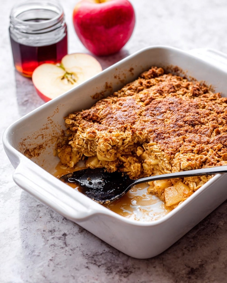 A white square dish holds a thick, golden brown baked oat casserole with a rough, crumbly texture on top. One large piece is missing, showing the inside layers of soft oats mixed with chunks of light brown baked apples. A black spoon rests inside the dish where the piece is taken out. Behind the dish, a red apple cut in half and a small jar of dark syrup sit on a white marbled surface. The scene has a warm and cozy feel, focusing on the hearty baked oats. photo taken with an iphone --ar 4:5 --v 7
