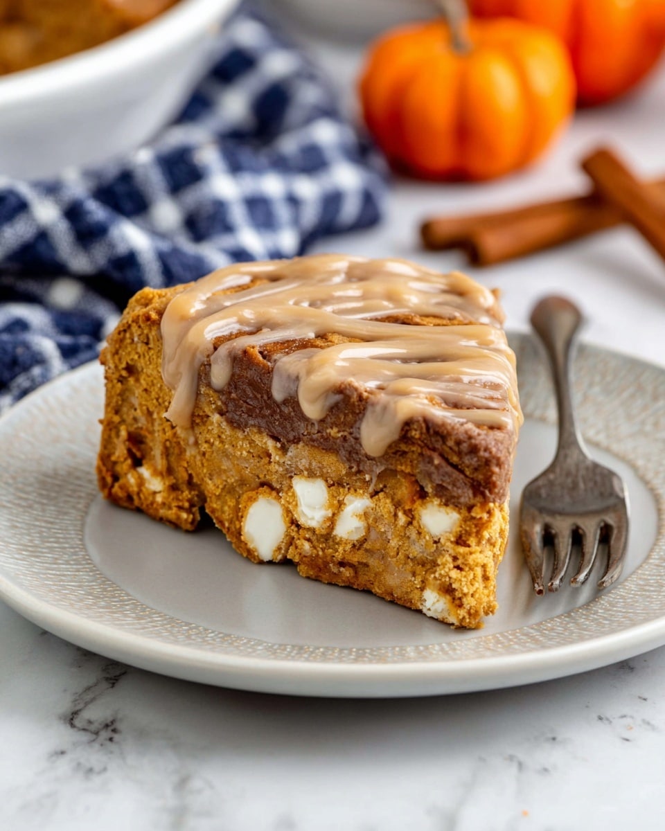 A slice of cake with two visible layers sits on a white plate with a light raised pattern around the edge, placed on a white marbled surface. The bottom layer is dense and moist with a golden brown color and chunks of white chocolate or cream inside. The top layer is thicker, a darker brown with a slightly cracked texture, and is covered by a light brown drizzle sauce in a swirl pattern. A vintage metal fork is resting on the right side of the plate, and a small orange pumpkin decoration along with a cinnamon stick are blurred in the background near a blue and white checkered cloth. Photo taken with an iphone --ar 4:5 --v 7