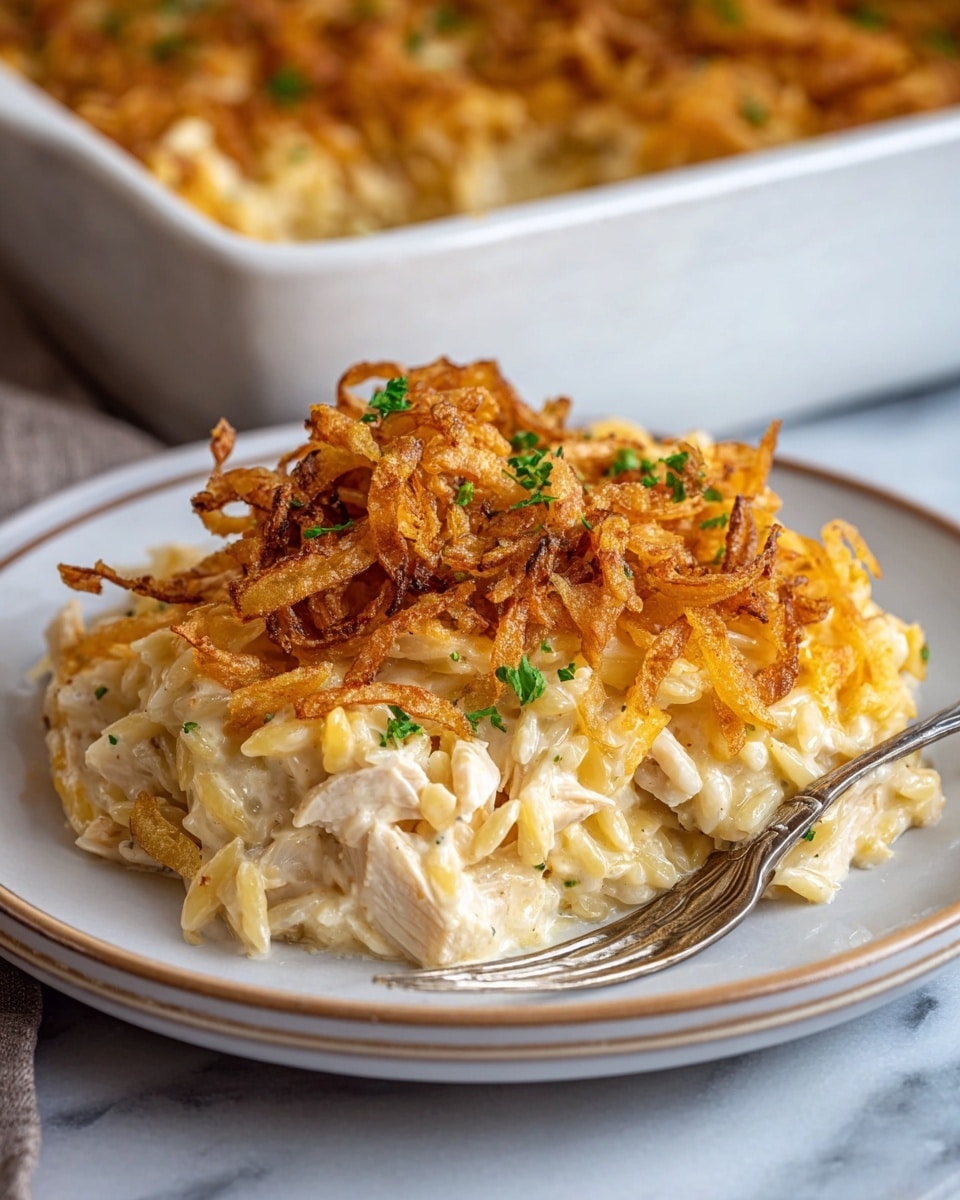 A white plate on a white marbled surface holds a single serving of creamy casserole with three visible layers. The bottom layer is composed of tender white chicken pieces mixed with soft, yellow-beige orzo pasta. The middle layer is a thick, smooth cream sauce that coats the chicken and orzo, giving a glossy texture. The top layer features a golden-brown pile of crispy fried onions scattered unevenly, sprinkled with small bits of green herbs for color contrast. A fork rests on the right side of the plate, partially embedded in the casserole. In the background, a white casserole dish filled with more of the same dish is blurred out. photo taken with an iphone --ar 4:5 --v 7