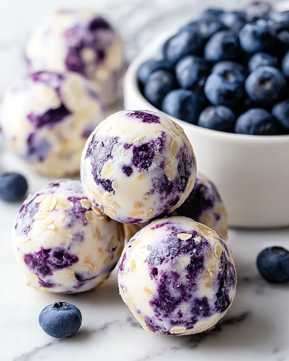The image shows several round snack balls placed on a white marbled surface. Each ball has two visual layers: a creamy white layer that looks smooth and slightly shiny, mixed with light beige small flakes, and patches of deep purple from embedded blueberries, giving a marbled effect. In the background, a white bowl filled with fresh, plump blueberries is partially visible. The overall look is fresh, soft, and appetizing with the contrast between the creamy white, light beige, and rich purple colors. Photo taken with an iphone --ar 4:5 --v 7