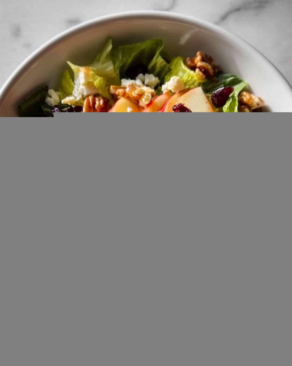 A bowl of fresh salad with several layers: the bottom layer consists of green leafy lettuce with a slightly rough texture; on top of this are thin slices of red-skinned apple, arranged in a loose circle; scattered over the apples are small pieces of white crumbled cheese with a soft texture; dark red dried cranberries are spread across the salad, adding depth in color and texture; whole walnut halves with a brown, wrinkled surface are also scattered on top; the entire salad has a light drizzle of golden honey or dressing giving a slight shine to the ingredients. The bowl is white, placed on a white marbled surface. photo taken with an iphone --ar 4:5 --v 7