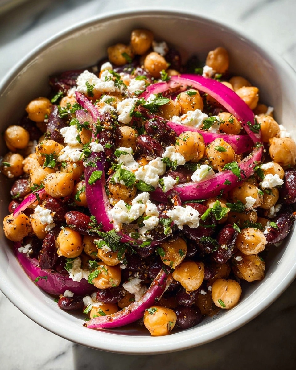 A close-up view of a white bowl filled with a vibrant salad made of three main layers: a base layer of golden chickpeas and dark red kidney beans, a middle layer of bright pink-red onion slices with a slight shine, and a top layer of small white crumbled cheese and chopped fresh green herbs scattered evenly. The salad looks mixed with a light coating of oil and black pepper specks, making the chickpeas and beans glisten. The bowl sits on a surface with a white marbled texture, and natural light highlights the fresh, colorful ingredients. photo taken with an iphone --ar 4:5 --v 7