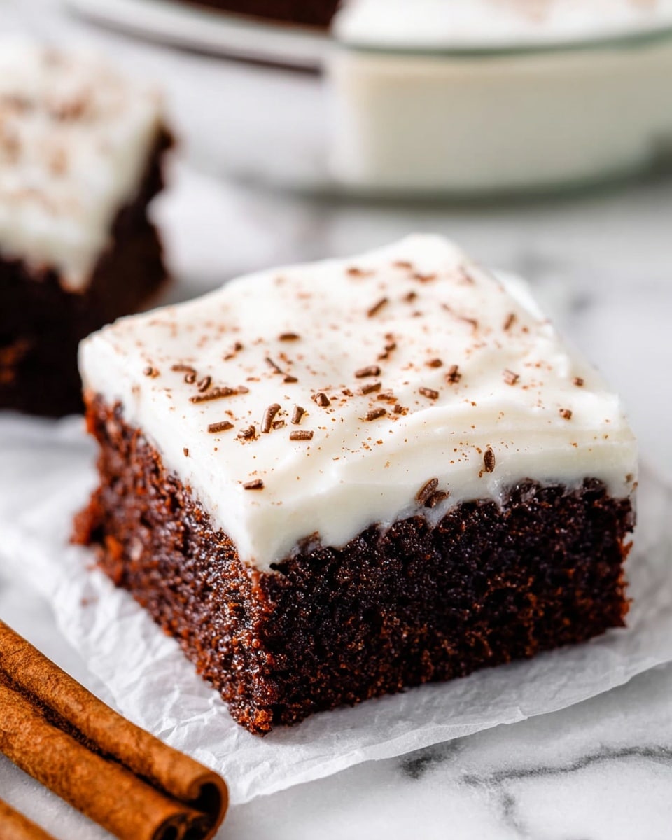 Two square chocolate brownies are stacked on top of each other on a white marbled surface. Each brownie has a thick, creamy white frosting layer on top, sprinkled lightly with brown powder, likely cinnamon or cocoa. The texture of the brownies looks dense and moist with a rich dark brown color. A woman's hand is holding the top brownie gently from the side, lifting it slightly. The background is soft and blurred with warm tones, showing faint shapes that hint at cinnamon sticks. Photo taken with an iphone --ar 4:5 --v 7