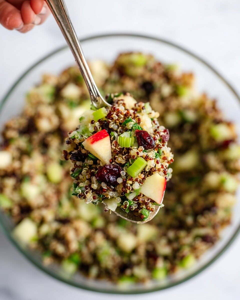 A close-up of a spoonful of a colorful quinoa salad held above a large clear glass bowl filled with the same salad, featuring three main layers: the base layer is light beige and fluffy quinoa mixed with darker quinoa grains, the middle layer contains bright green chopped celery and herbs, and the top layer includes small red apple cubes and dried dark brown cranberries scattered throughout; the texture is a mix of smooth quinoa and crunchy vegetable bits, set against a white marbled background, with a woman's hand holding the spoon. photo taken with an iphone --ar 4:5 --v 7