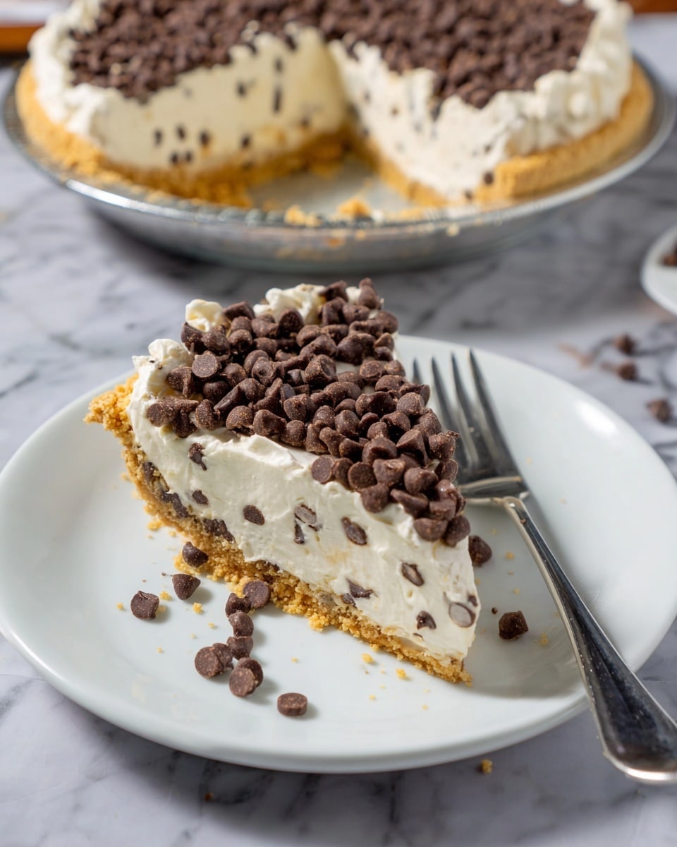 A slice of pie on a white plate shows three main layers: a golden brown crumbly crust at the bottom, a thick creamy off-white filling with small chocolate chips mixed inside, and a topping of whipped cream covered with a thick layer of dark brown mini chocolate chips. The top layer is textured with scattered chocolate chips, some falling onto the plate. A silver fork lies next to the slice on the plate, all placed on a white marbled surface. In the background, the rest of the pie on a white dish shows the same layers. photo taken with an iphone --ar 4:5 --v 7