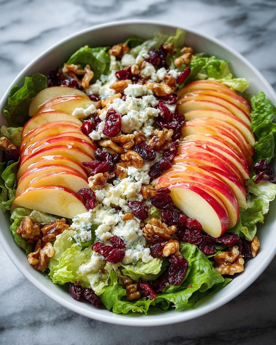 A white bowl filled with a fresh salad is shown on a white marbled surface. The salad has three main layers: the bottom layer is green leaves of lettuce, shiny and fresh; the middle layer consists of thin slices of red and yellow apple, arranged in fan shapes on top of the lettuce; the top layer is a mix of white crumbled cheese, bright red dried cranberries, and golden-brown walnuts evenly spread over the salad. The salad is lightly dressed, giving the apples a glossy look. photo taken with an iphone --ar 4:5 --v 7