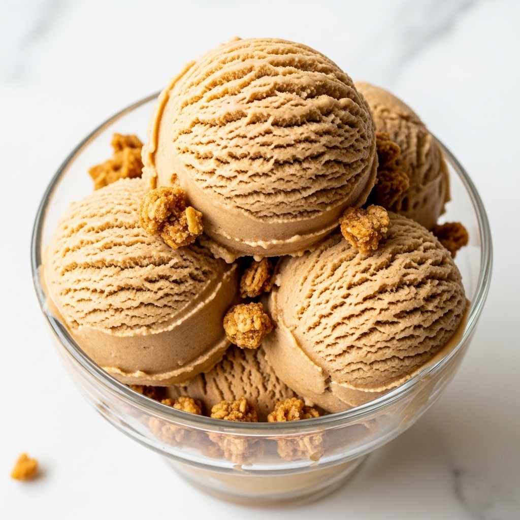 A close-up view of a transparent glass dessert cup filled with three smooth, light brown ice cream scoops forming the base and one larger, textured scoop of the same flavor on top. The ice cream is surrounded and topped with small pieces of crunchy granola that add a rough, golden-brown contrast to the creamy texture. The cup sits on a white marbled surface, and natural light softly highlights the ice cream's creamy texture and subtle ridges. photo taken with an iphone --ar 4:5 --v 7