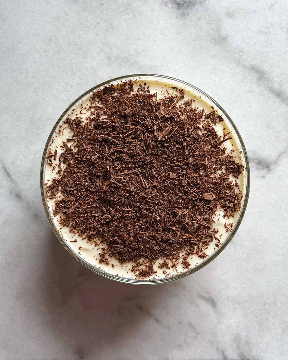 A round glass bowl filled with one visible layer of creamy light beige dessert, topped with a generous, uneven layer of finely grated dark brown chocolate shavings that cover almost the entire surface. The bowl is set on a white marbled textured surface, with soft natural lighting highlighting the smoothness of the cream and the delicate texture of the chocolate on top. photo taken with an iphone --ar 4:5 --v 7