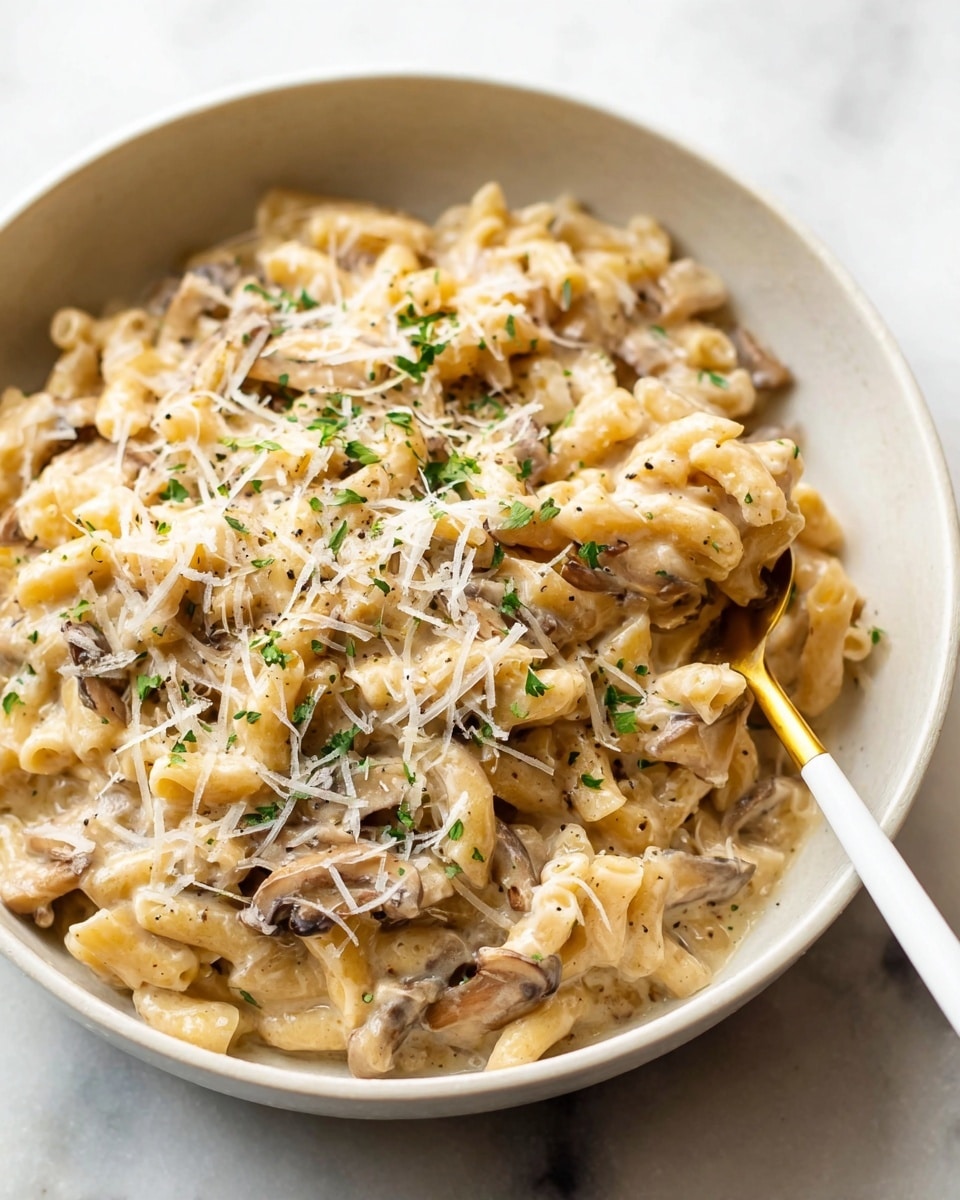A creamy, light beige pasta dish is served in a white bowl, featuring small, short tubular pasta mixed with thinly sliced mushrooms coated in a thick sauce. The pasta and mushrooms have a glossy texture, with visible flecks of black pepper and finely chopped green herbs sprinkled on top. Shredded white cheese lightly covers the surface, adding a soft texture. A golden spoon with a white handle holds a small bite of the pasta, rising slightly from the bowl, all placed on a white marbled surface. photo taken with an iphone --ar 4:5 --v 7