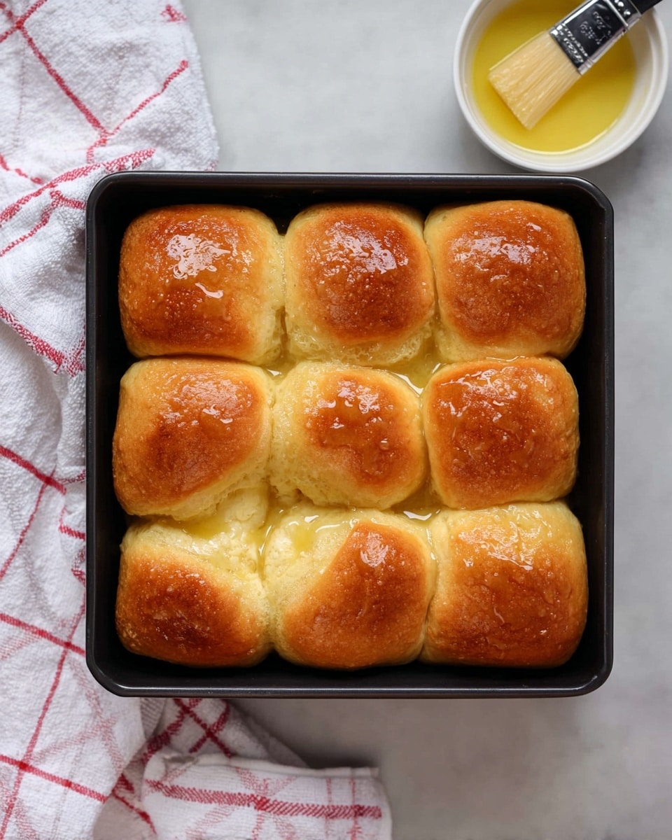 The image shows a close-up of a black baking pan filled with nine soft, golden brown bread rolls, arranged in a 3x3 grid. Each roll has a smooth, slightly uneven surface with a warm toasted color on top. One roll is being lifted above the rest by a woman's hand, revealing its inside texture which is light, fluffy, and pale yellow with small air holes throughout. The background is a white marbled texture. photo taken with an iphone --ar 4:5 --v 7
