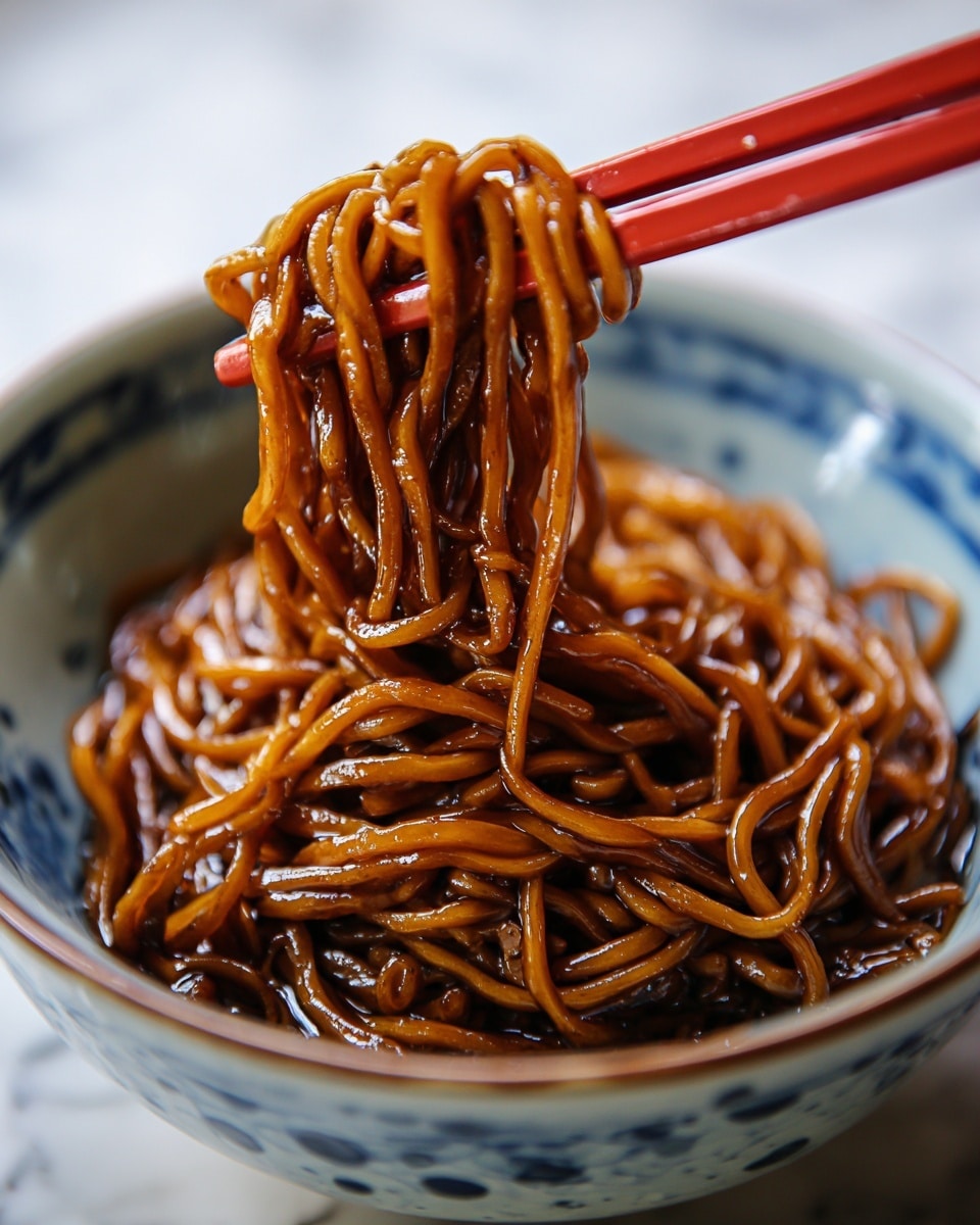 A deep white bowl filled with a single layer of glossy, dark brown noodles coated evenly in a thick sauce, creating a rich shine on their smooth surface. Bright red chopsticks hold a small twirl of noodles lifted from the bowl, showing the detailed texture of each strand. The bowl sits on a white marbled surface that adds a clean, subtle contrast to the warm tones of the noodles. photo taken with an iphone --ar 4:5 --v 7