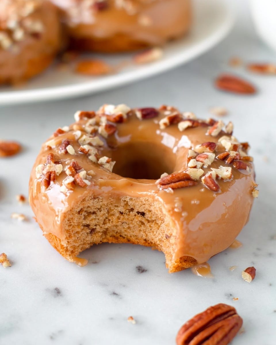 The image shows a single donut with a bite taken out of it, placed on a white marbled surface. The donut has one main layer of light brown dough with a soft, moist texture visible in the bitten area. The donut is fully covered in a shiny caramel-colored glaze that adds a smooth surface on top. Scattered pieces of chopped pecans are sprinkled unevenly over the glaze, adding a crunchy texture and darker brown color contrast. Around the donut on the white marbled surface, there are a few loose pecan halves. In the background, another white plate is slightly visible, holding more donuts of the same kind. photo taken with an iphone --ar 4:5 --v 7