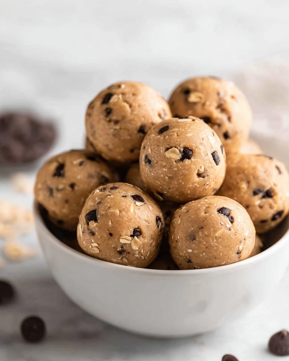A white bowl is filled with about fourteen round energy balls, each one light brown with small dark chocolate chips evenly spread throughout, showing a slightly rough texture with oats visible in the mixture. The balls are piled on top of each other, filling the bowl to the edge, with some balls closer to the camera and others stacked behind. The background features a white marbled texture with scattered chocolate chips blurred softly, adding depth to the image. The lighting is soft and natural, highlighting the shiny, slightly sticky surface of the energy balls. photo taken with an iphone --ar 4:5 --v 7