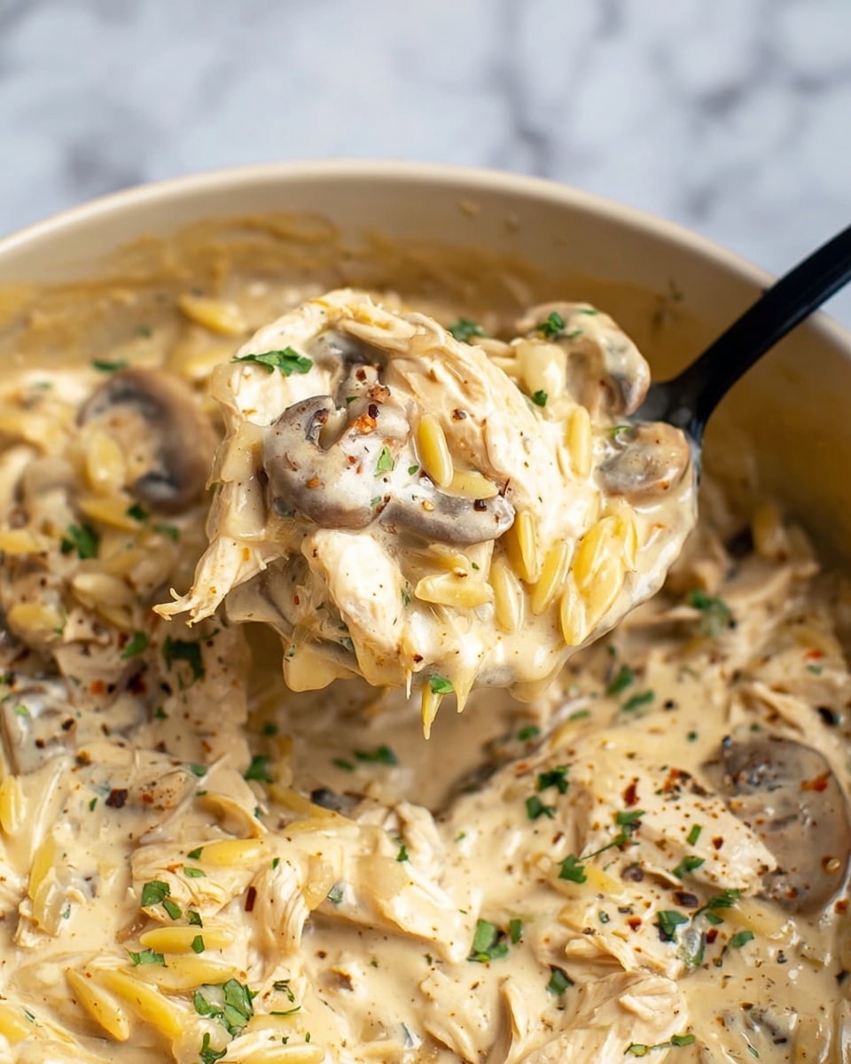 The image shows a close-up of a creamy dish with multiple layers and textures. At the base, there are smooth, thick beige cream sauce layers mixed with tender, slightly shredded white chicken pieces. Scattered throughout are small, oval-shaped light yellow orzo pasta and sliced brown mushrooms with soft edges. The dish is speckled with finely chopped green herbs and small black pepper flakes, adding subtle color contrast. A black spoon holds a scoop of the creamy mixture, lifting a portion with orzo, chicken, and mushroom pieces, all coated in the rich cream sauce. The background is a white marbled texture. Photo taken with an iphone --ar 4:5 --v 7
