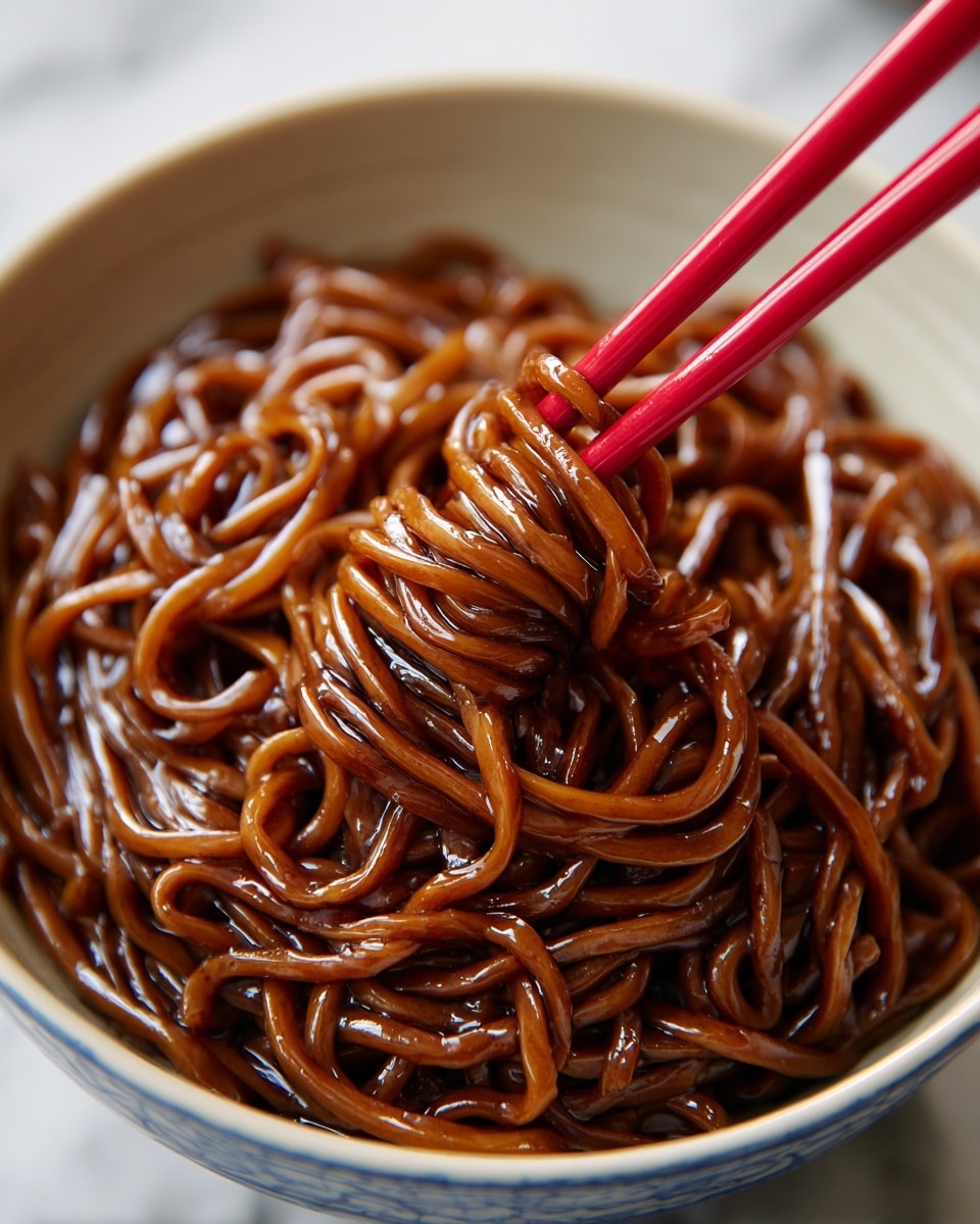 A close-up of dark brown noodles coated in a thick, glossy sauce, lifted by bright red chopsticks above a deep white bowl with blue speckled patterns. The noodles are twisted and shiny, sitting in a pool of the rich sauce at the bottom of the bowl. The bowl rests on a white marbled surface, with the focus on the noodles and chopsticks showing a textured, smooth, and slightly oily look. photo taken with an iphone --ar 4:5 --v 7
