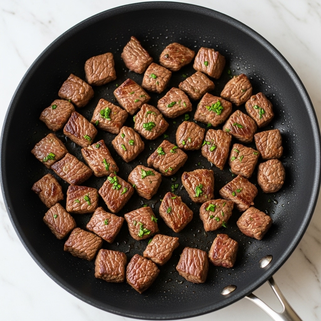 This image shows a black frying pan filled with small cubed pieces of cooked beef, each piece having a rich brown sear and a slightly glossy texture from the cooking juices. The beef cubes are scattered evenly across the pan with a sprinkling of finely chopped fresh green herbs on top, adding a touch of brightness. The pan itself rests on a white marbled surface, with the lighting highlighting the juicy and tender look of the meat. photo taken with an iphone --ar 4:5 --v 7