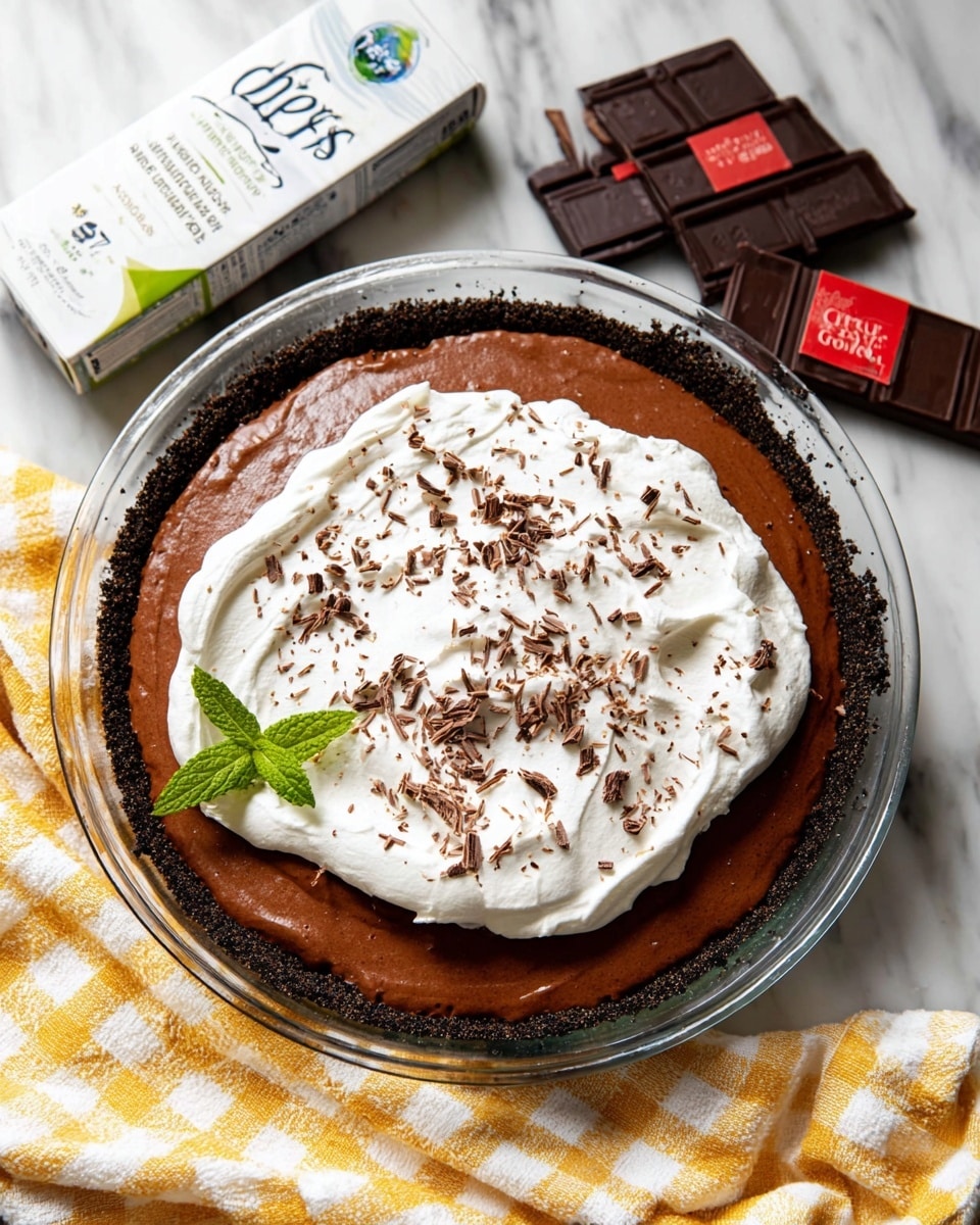 The image shows a pie with three visible layers in a clear glass pie dish on a white marbled surface. The bottom layer is a dark, crumbly cookie crust, thick and holding the pie together. The middle layer is a smooth, rich chocolate filling that is glossy and evenly spread. The top layer is a fluffy, white whipped cream, spread loosely with soft peaks and sprinkled with small chocolate shavings. On one side of the whipped cream, there is a small green mint leaf for decoration. Next to the pie is a white and yellow checkered cloth, a clear egg carton, and two bars of dark chocolate with red accents on their wrappers. Photo taken with an iphone --ar 4:5 --v 7